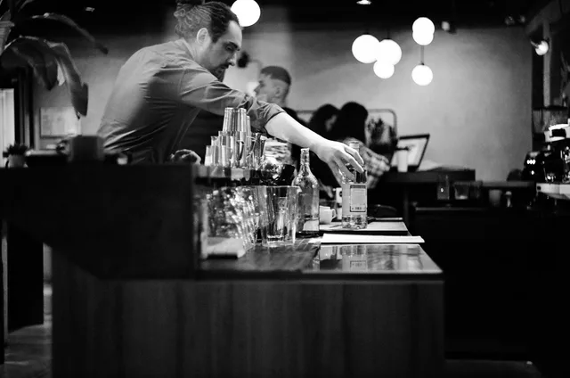 A monochrome image capturing a bartender arranging bottles behind a bar counter, creating a classic atmosphere. Tillstånd och regler för krogdrift: så håller du alkohol, livsmedel och skatt rätt